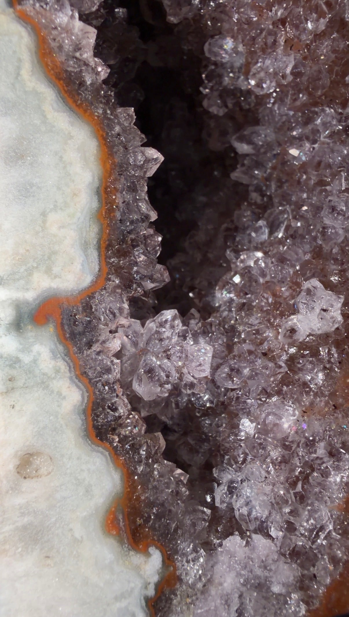 Close-up of a geode with translucent lavender-purple amethyst crystals, a dark central cavity, and a white-to-gray banded outer layer with rust-colored edges.
