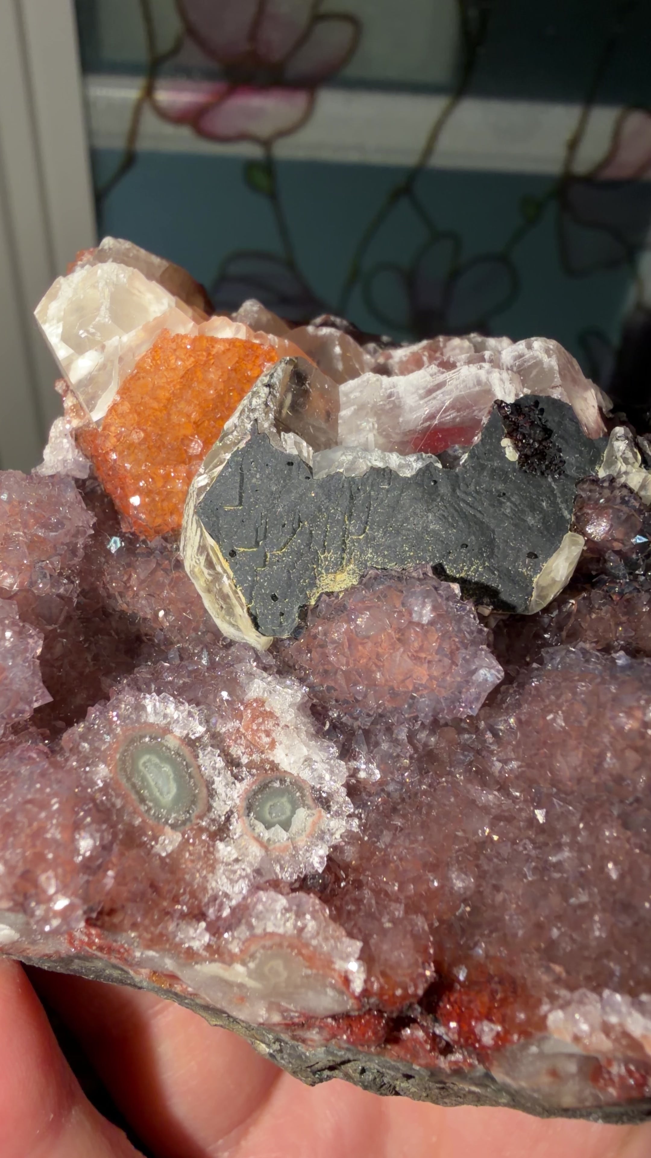 Close-up of a multi-mineral specimen showing its purple stalactite, orange calcite, red and black hematite formations. 