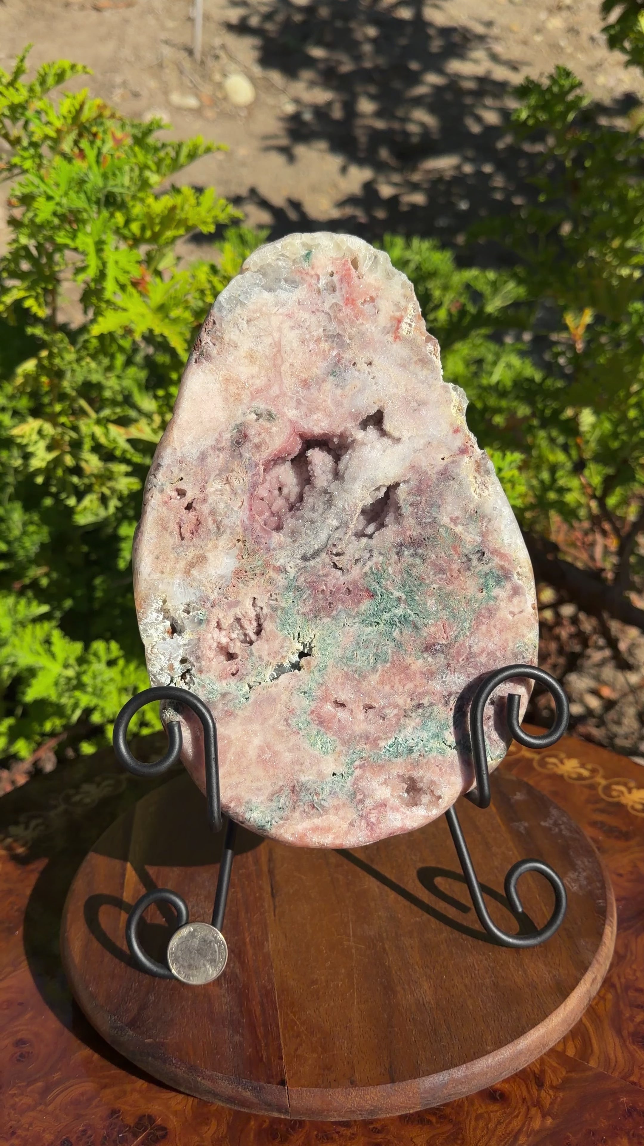 Large egg shaped Brazilian pink amethyst plate displayed on black metal easel. Green mossy features al throughout. Botryoidal formations. 