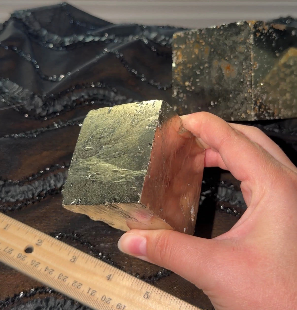 A hand holding a gold/bronze metallic pyrite cube corner. You can see the fingers in the reflection of the surface.